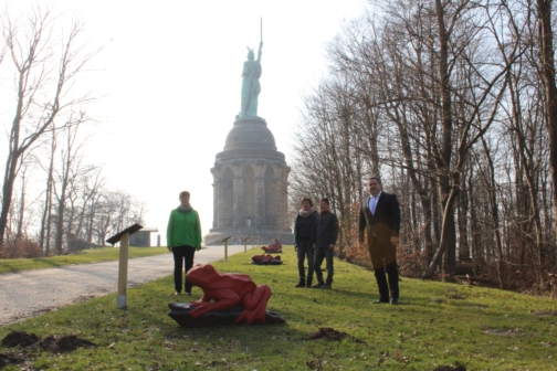 Rot geschnitzter Frosch und andere Tiere vor dem Hermannsdenkmal mit vier präsentierenden Leuten